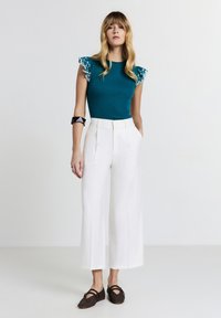 Top teal a costine con maniche a volants in pizzo, abbinato a pantaloni bianchi a vita alta e a gamba larga e scarpe ballerine in pelle marrone scuro con texture.