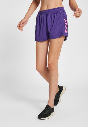 Slank person iført lilla atletikshorts med lyserøde chevroner og sorte løbesko, står med den ene fod løftet.