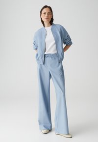 Veste bomber bleu clair avec fermeture éclair à l'avant et manches retroussées, assortie à un pantalon large avec taille élastique. Chemise blanche en dessous.