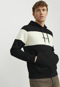 Sort og creme farveblokket hættetrøje med snøre, frontlomme og logo på brystet. Blødt stof, afslappet pasform.