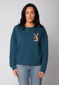 Femme portant un sweat-shirt bleu-vert avec un petit design de carte de tarot intitulé "Les Amants" sur le haut à gauche de la poitrine, associé à un jean bleu clair.