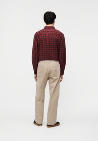 Άνδρας που στέκεται και κοιτάζει προς τα πίσω φορώντας κόκκινο και μαύρο καρό πουκάμισο, μπεζ παντελόνι με πίσω τσέπες και μαύρα αθλητικά παπούτσια με καουτσούκ σόλες.