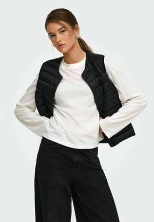 Jeune femme aux cheveux bruns attachés en queue de cheval, portant une chemise blanche à manches longues, un gilet matelassé noir et un pantalon noir sur un fond uni.