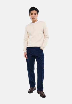 Homme debout, une main dans la poche, portant une chemise henley beige à manches longues, un pantalon bleu marine et des chaussures marron foncé sur fond blanc.