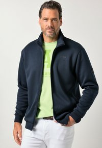 Donkerblauwe sweatshirt met rits en hoge kraag, voorzien van een subtiel logo op de borst, gecombineerd met een lichtgroen T-shirt eronder.