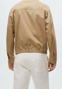 Brun, let jakke med en glat tekstur, elastisk talje og lange ærmer, parret med lyse bukser. Simpelt design.