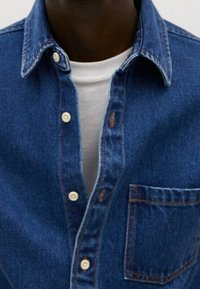 Donkerhuidige persoon draagt een ongeknopte blauwe spijkerblouse over een witte T-shirt met ronde hals.