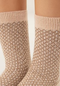 Gambe con calzini beige lavorati a maglia con fili metallici dorati e bianchi, polsini a coste e motivo strutturato su sfondo chiaro.