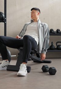 Siva športna jakna s črnimi črtami, bela majica, črne hlače in bele teniske. Hladilnice in fitnes oprema v ozadju.