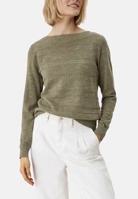 Pull tricoté vert olive avec un col large, présentant des rayures horizontales et des détails en œillets ; associé à un pantalon blanc taille haute.