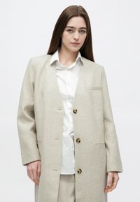 Jasnobeżowa marynarka z mieszanki wełny o luźnym kroju, z pojedynczą kieszenią, dwoma guzikami i klapą z wycięciem, noszona na białej koszuli.