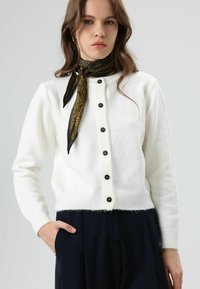 Cardigan blanc en tricot avec boutons noirs, ourlet côtelé et manches longues. Porté avec une écharpe noire et dorée à motifs pour plus de détails.