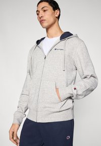 Sweat à capuche gris avec fermeture éclair, capuche doublée en bleu marine, poches avant et logo sur la poitrine. Confectionné en tissu doux, avec poignets et ourlet côtelés.