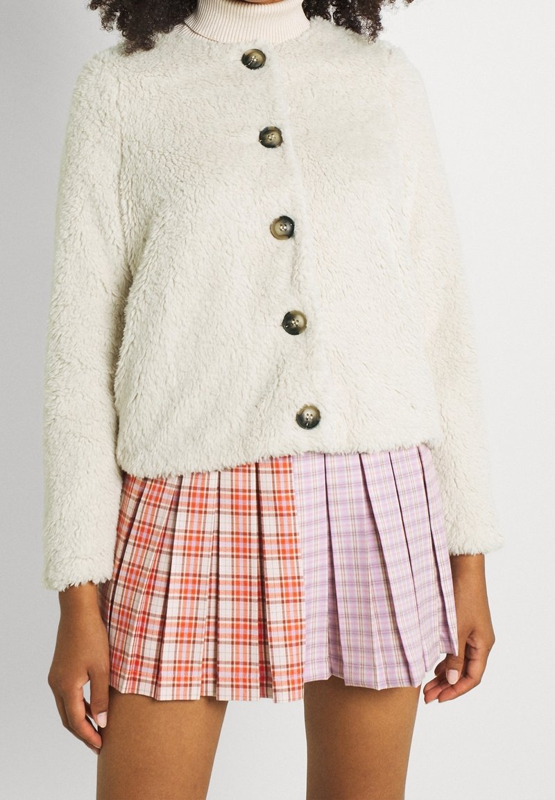 Femme portant une veste crème duveteuse boutonnée sur une mini-jupe plissée avec des panneaux à carreaux orange et lavande.
