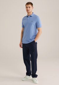 Camiseta polo de manga corta azul claro con un ajuste slim, detalle del logo negro en el pecho, combinada con pantalones oscuros azul marino y zapatillas blancas.
