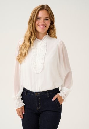 Witte blouse met gerimpelde voorkant en een hoge hals, lange doorschijnende mouwen met gerimpelde manchetten, gecombineerd met donkere spijkerbroeken.
