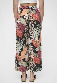 Blomstrete vidbenbukser i svart stoff, med store oransje og grønne blader, små blomster, og snøring i midjen. Sandaler synlige.