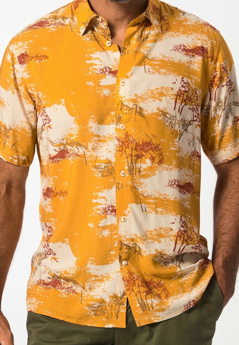 Chemise à manches courtes boutonnée avec des motifs abstraits de paysage jaunes et beiges portée par une personne les mains dans les poches.
