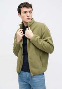 Olivovo zelená fleece bunda s vysokým golierom, predným zipsom a bočnými vreckami, nosená cez tmavé tričko, v kombinácii so svetlomodrými džínsami.