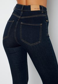 Tmavě modré úzké džíny s žlutým šitím, vysokým pasem a koženým štítkem na zadní straně s názvem značky a logem.