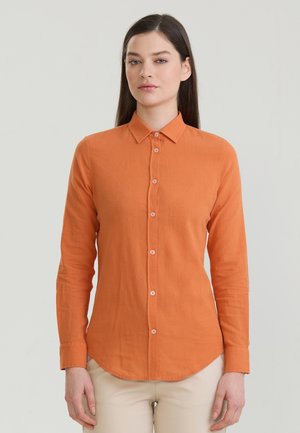 Naine pikkade tumedate juustega, kes kannab keha ümberistuvat oranži triiksärki pikkade varrukatega ja beežid pükse, seisab ühevärvilise heleda tausta ees.