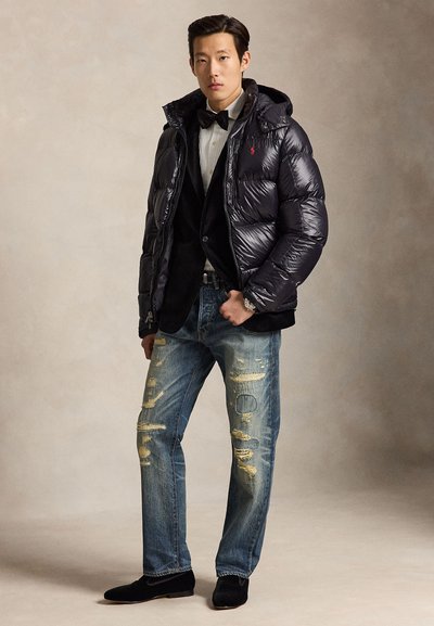 Mann trägt eine glänzende schwarze Daunenjacke über einem schwarzen Blazer, weißem Hemd mit Schleife, abgenutzten blauen Jeans und schwarzen Schuhen, steht vor einem schlichten Hintergrund.