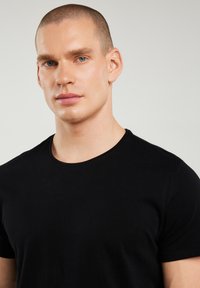 Lühikeste juustega mees siniste silmadega, seljas lihtne must ümara kaelusega särk, helehall taust.