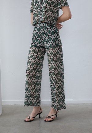 Femme portant un pantalon et un haut assortis à motifs circulaires verts et beiges, associés à des sandales à talons noires à brides.