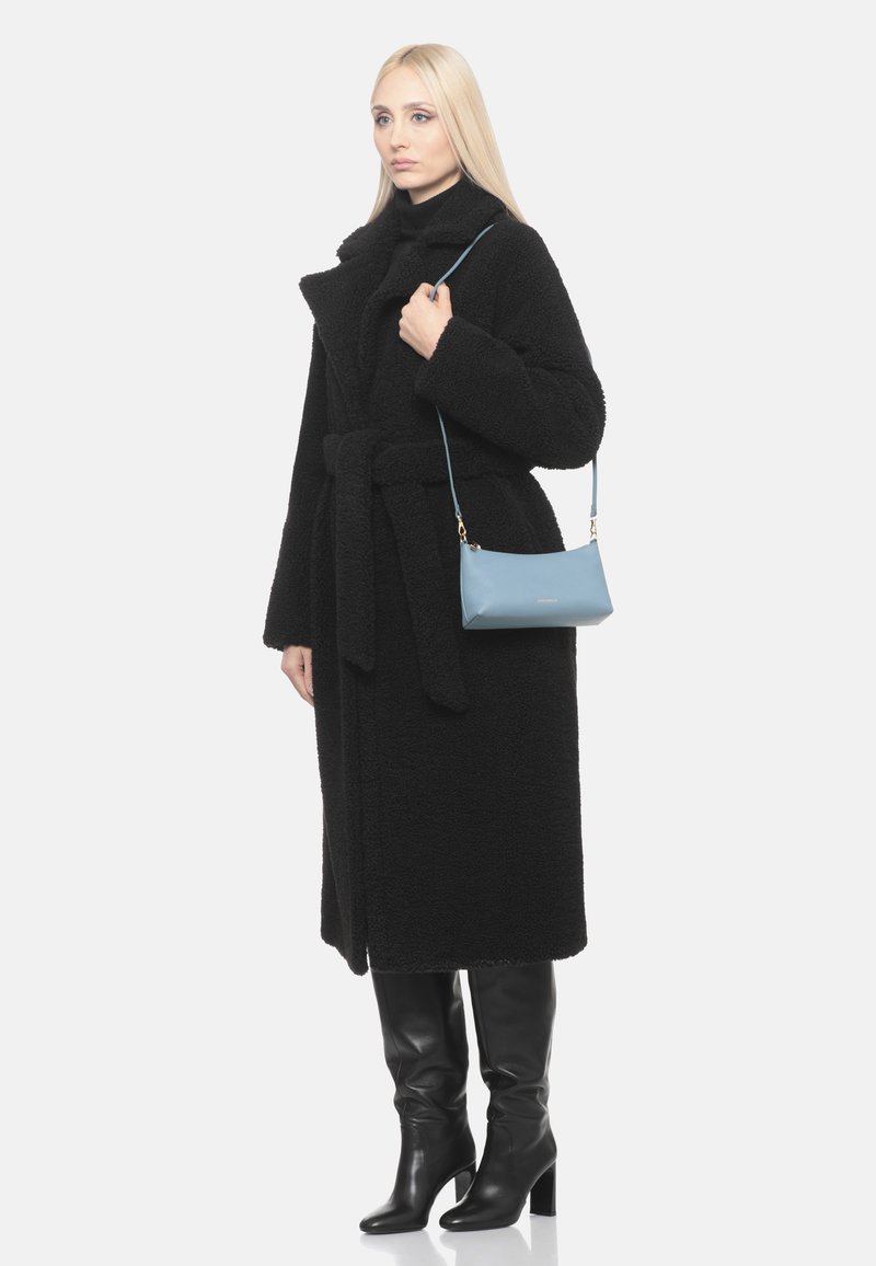 Donna bionda che indossa un lungo cappotto nero testurizzato e stivali neri con tacco, con una piccola borsa azzurro chiaro sulla spalla.
