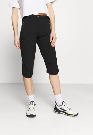 Woman wearing black knee-length pants with zipper pockets, white crew socks, and white athletic shoes with black and neon details.