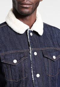 Denim jacket with a cream faux fur collar, dark blue fabric, two chest pockets, and silver snap buttons along the front and pockets.