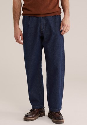 Muž v tmavomodrých voľne strihaných džínsoch, hnedých textúrovaných topánkach a hrdzavej mikine, stojaci na jednofarebnom béžovom pozadí.