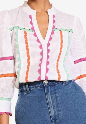 Witte blouse met knopen en een kleurrijke geschulpte rand in roze, groen en oranje, gecombineerd met een high-waist blauwe spijkerbroek.