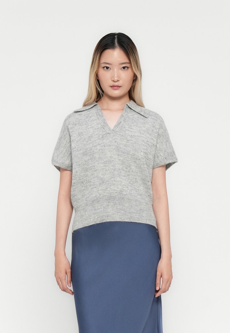Giovane donna con lunghe capelli ombre che indossa un maglione grigio a maglia a maniche corte con colletto e una gonna blu in raso, in piedi contro uno sfondo bianco.