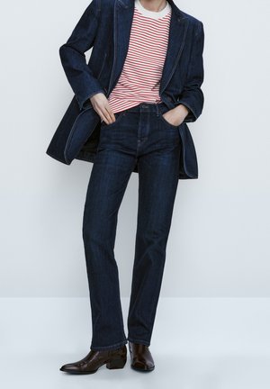 Person iført mørkeblå denimjakke og jeans, rød- og hvidstribet skjorte og mørkebrune ankelstøvler, hænderne i lommerne.