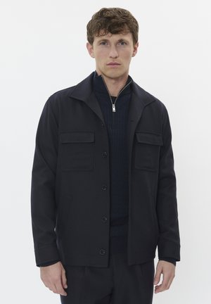 Marineblaue Jacke mit einer glatten Textur, ausgestattet mit einem Kragen, einer Knopfleiste und zwei Fronttaschen. Getragen über einem dunklen Pullover.
