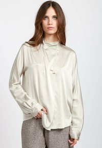 Blusa di seta color beige chiaro con colletto alto, piega frontale e dettaglio con nappina. Le maniche lunghe presentano polsini con bottoni e una vestibilità comoda.