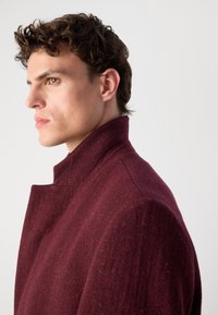 Sacou din lână burgundy cu guler înalt și țesătură texturată. Are o siluetă ajustată și detalii de cusătură vizibile pe umeri.