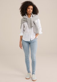 Hellgrauer, gestrickter Pullover, um die Schultern gebunden, weißes Hemd mit Knöpfen, hellblaue Jeans und leichte Sneakers mit strukturierten Sohlen.