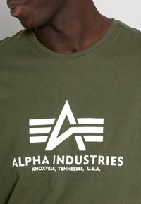 Olivengrünes Baumwoll-T-Shirt mit einem großen weißen Logo mit Streifen und einem "A". Rundhalsausschnitt und kurze Ärmel. Schlichtes, modernes Design.
