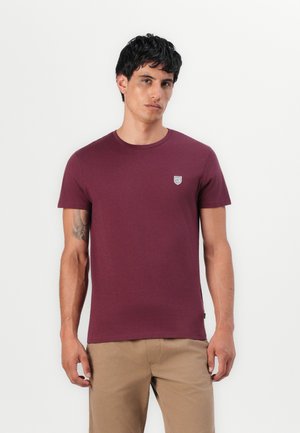 T-shirt à manches courtes marron, fabriqué en tissu doux ; présente un petit emblème sur la poitrine gauche et un col rond. Design simple, taille standard.