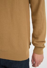Jersey de punto en marrón claro con una superficie texturizada, puños y dobladillo acanalados. Presenta un diseño moderno y casual, apto para llevar en capas.