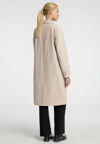 Manteau beige clair au design ajusté, avec une fente unique et des détails de poignets à trois boutons, associé à un pantalon noir à jambes larges.