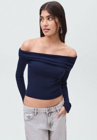 Námořnická modrá žebrovaná halenka s odhalenými rameny, dlouhými rukávy, přiléhavým střihem a texturovanou tkaninou. Kombinováno se světle šedými džíny s vysokým pasem.