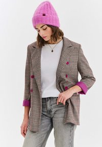 Kárované sako v hnedej a krémovej farbe s ružovými gombíkmi a detailmi na manžetách, doplnené o biely top a sivé rifle. Modelka má na hlave ružovú pletenú čiapku.