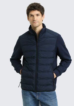 Homme aux cheveux courts et foncés portant une veste matelassée bleu marine à fermeture éclair et un jean bleu, debout les mains dans les poches devant un fond uni.