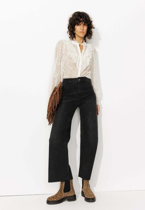 Blouse en dentelle blanche avec un col à volants, jeans noirs à taille haute et jambes évasées, et bottines chevilles en imprimé léopard marron. Sac à franges marron.