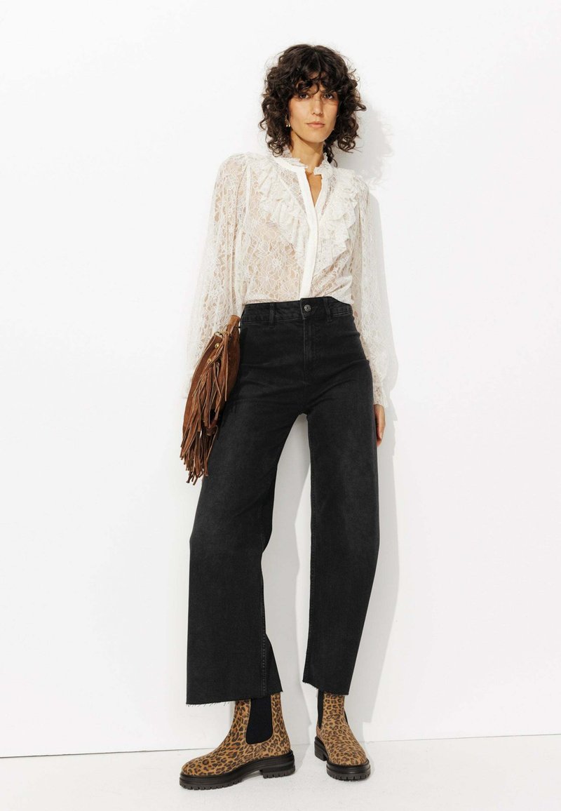 Blouse en dentelle blanche avec un col à volants, jeans noirs à taille haute et jambes évasées, et bottines chevilles en imprimé léopard marron. Sac à franges marron.