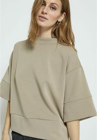 Béžový oversized top s vysokým výstřihem, krátkými dolman rukávy a hladkým povrchem; má široký lem na spodním okraji.