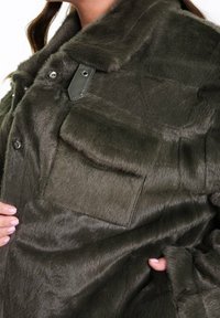 Olivově zelená umělá kožešinová bunda s jemnou texturou, špičatým límcem, patentkami a náprsní kapsou. Vyznačuje se elegantním designem a hladkým povrchem.